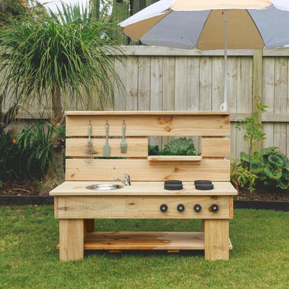 Wooden outdoor kitchen setup with grill, sink, and utensils on a grassy area with cacti in the background in papamoa